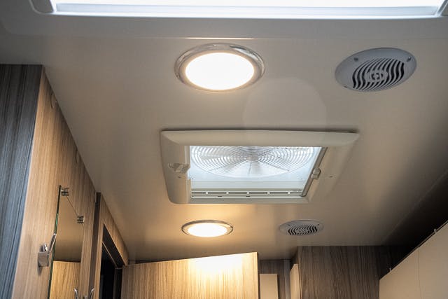 The image shows the interior ceiling of a bathroom in a 2019 Benimar Tessoro T486. It includes a central vent with a fan, two circular lights, and two small circular vents. The ceiling and walls are made of wood and light-colored materials.