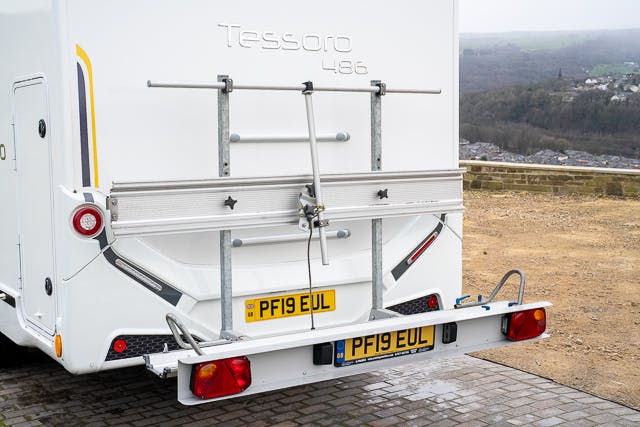 The image shows the rear end of a white 2019 Benimar Tessoro T486 motorhome with a number plate "PF19 EUL." Attached to the back is a bicycle rack. The motorhome, labeled "Tessoro 486," is parked on a paved area with a scenic landscape in the background.