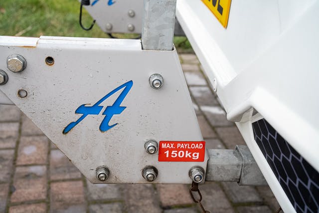 Close-up of a white metal component of the 2019 Benimar Tessoro T486 with bolts and a blue letter "A" logo. A red and white label indicates a maximum payload of 150 kg. The vehicle part is situated on a paved surface with grass visible in the background.