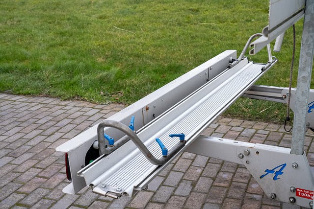 An aluminum ramp attached to a 2019 Benimar Tessoro T486 trailer is positioned over a paved surface with grass in the background. The ramp includes blue handle grips and a notched metal support arm on the side.