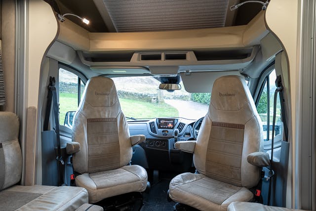 This image shows the interior of a 2019 Benimar Tessoro T486 motorhome from the midsection, focusing on the driver and passenger seats. The seats are upholstered in beige fabric and have armrests. The dashboard and steering wheel are visible, along with a window view outside.