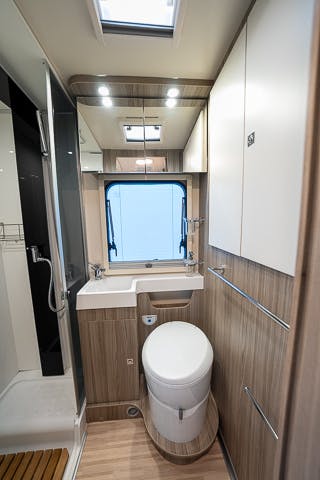 The image shows the interior of a compact and modern RV bathroom in the 2019 Benimar Tessoro T486. It features a toilet, a small sink, a window, a shower area with a glass door, and wooden cabinetry. The space is efficiently designed with overhead lighting and contemporary finishes.
