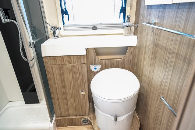A compact bathroom in the 2019 Benimar Tessoro T486 features a white toilet, a wooden cabinet with a sink and faucet, and a window above the sink. A towel rack is mounted on the wall, and the corner of a shower area with a glass door is visible on the left.