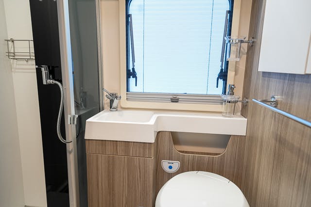 A compact bathroom in the 2019 Benimar Tessoro T486 features a rectangular sink set in a wooden vanity, with a toilet beneath and a large window above. To the left, there's a wall-mounted showerhead and a glass door leading to the shower area, with several shelves visible on the walls.