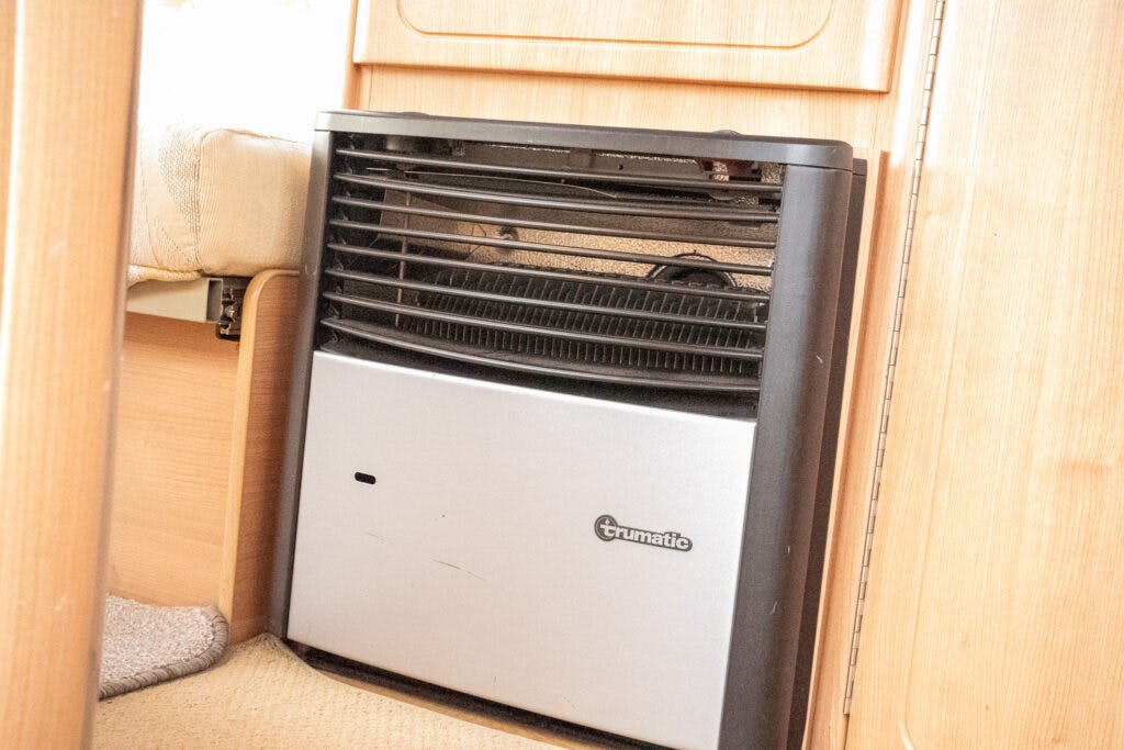 A Trumatic gas heater is installed on a wooden panel in a room, reminiscent of the sophisticated interior of a 2006 Auto-Sleepers Nuevo EK. The heater boasts a metallic gray front panel and a black grill above it. The floor is carpeted, and to the left of the heater, part of a beige couch is visible.