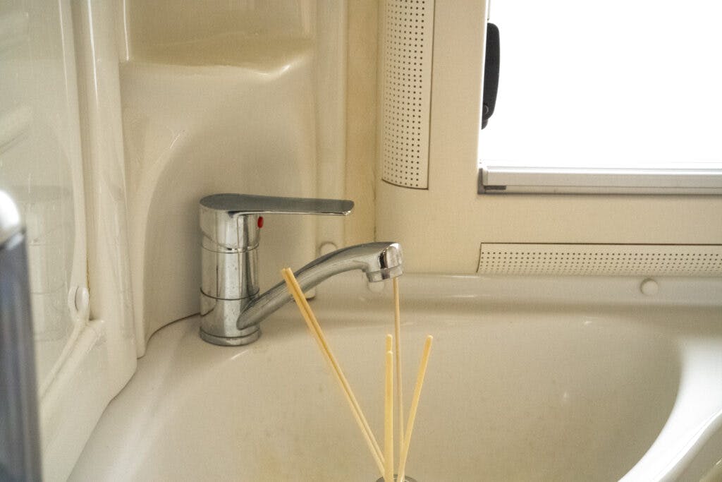 A chrome faucet is installed over a white ceramic sink. The faucet is positioned near a window with a beige frame, reminiscent of the clean design found in a 2006 Auto-Sleepers Nuevo EK. A set of bamboo skewers stands in a container on the sink’s surface, adding to the bright and natural feel of the area.