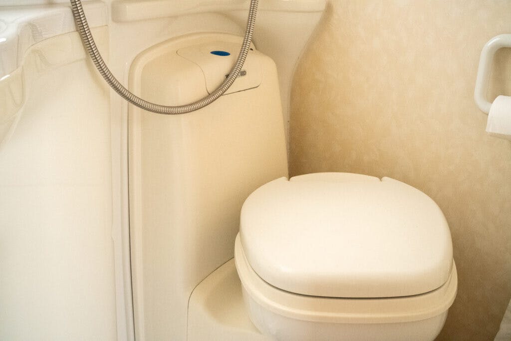 A compact bathroom in the 2006 Auto-Sleepers Nuevo EK features a cream-colored toilet with a closed lid. Attached to the side of the toilet is a flexible metal hose, possibly for a bidet or shower attachment. The background includes a light-colored, textured wall and a partial view of a bathtub.