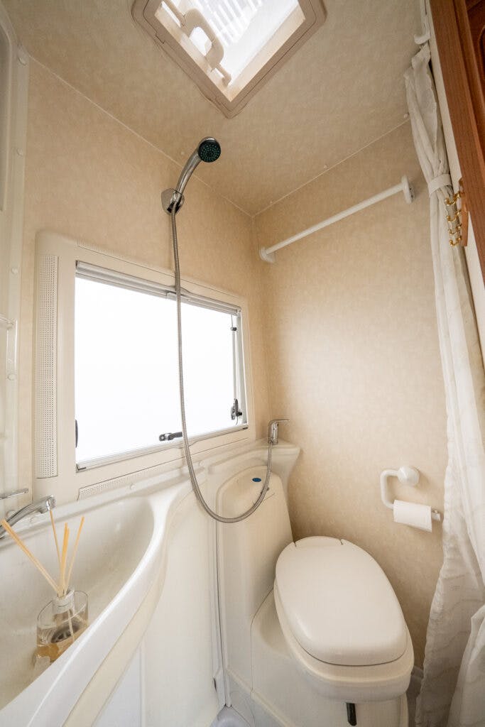The image shows a compact bathroom inside a 2006 Auto-Sleepers Nuevo EK, featuring a white toilet next to a small sink. A showerhead is mounted on the wall above the toilet. There is a window with a ledge, a white curtain on a rail, and a ceiling vent. A toilet paper holder is attached to the wall.