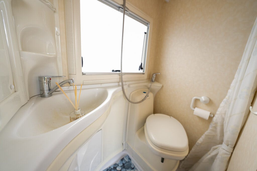 A compact bathroom in the 2006 Auto-Sleepers Nuevo EK features a white sink with a chrome faucet, a reed diffuser, and a small window above. Adjacent is a white toilet next to a wall-mounted toilet paper holder. A shower hose is affixed to the sink area. The floor has a blue pattern.