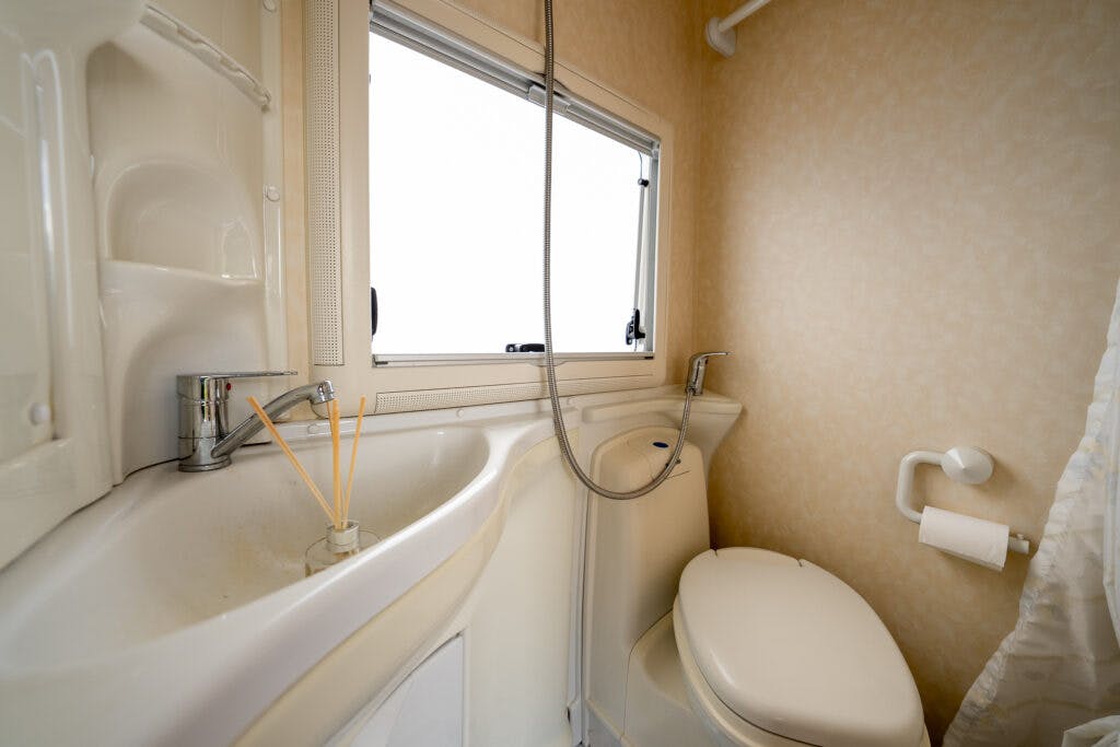A cozy bathroom in the 2006 Auto-Sleepers Nuevo EK features a sink with a faucet and a reed diffuser placed on it. Above the sink is a showerhead attached to a flexible hose. The space also includes a toilet with a wall-mounted holder for the toilet paper roll, all complemented by natural light from the window.