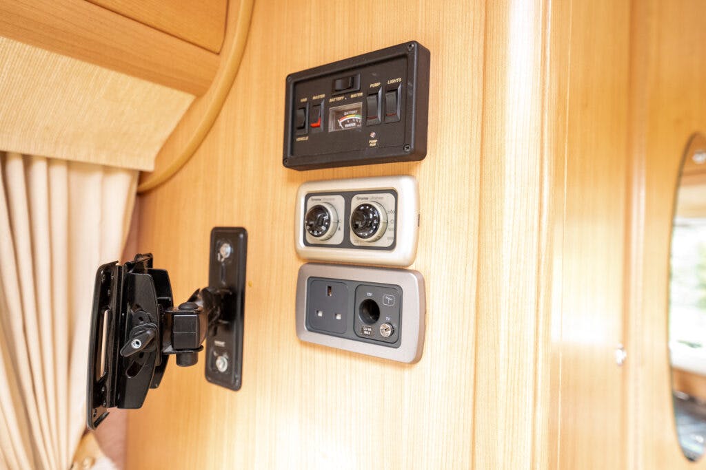 A close-up view of a wall panel in an RV featuring various control switches and outlets. The panel includes controls and power sockets, with curtains partially visible to the left and reflective wooden panels surrounding the area, typical of the 2006 Auto-Sleepers Nuevo EK.