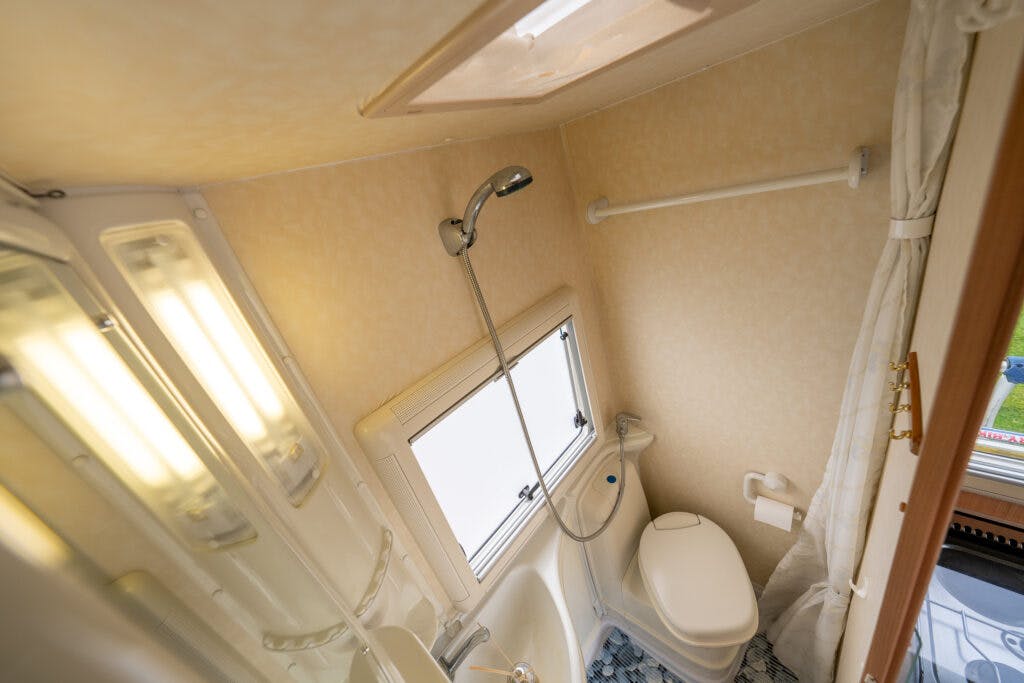 A compact bathroom with beige walls inside the 2006 Auto-Sleepers Nuevo EK. The room features a small shower area with a handheld showerhead, a window, and a white toilet. The sink is integrated into the shower unit, and there is a towel rack and a shower curtain.