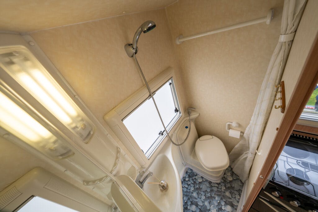 A compact RV bathroom in the 2006 Auto-Sleepers Nuevo EK features a showerhead attached above a small sink, a toilet adjacent to a window, and a towel rack. The space is lined with light-colored walls and a patterned blue floor, with a partially visible mirror and light fixture.