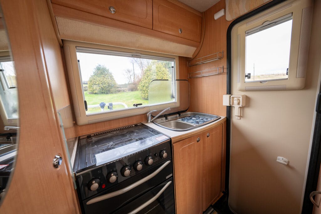 Interior of a small, compact kitchen inside a 2006 Auto-Sleepers Nuevo EK RV. It features a stovetop with an oven below, a small sink with a single faucet, and some cabinets for storage. A window above the sink provides natural light, and there is a shelf for small items.