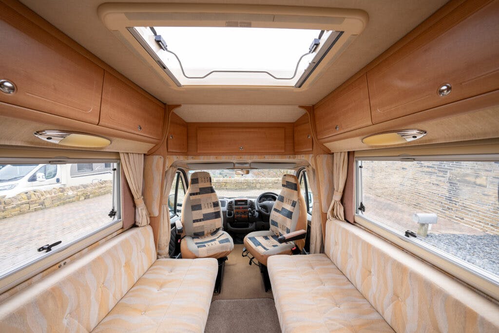 The interior of the 2006 Auto-Sleepers Nuevo EK camper van with beige upholstery features two front seats, cabinets above, large windows on both sides, and skylights above. The design is spacious with wood furnishings. Outside, other vehicles and brick structures are visible.