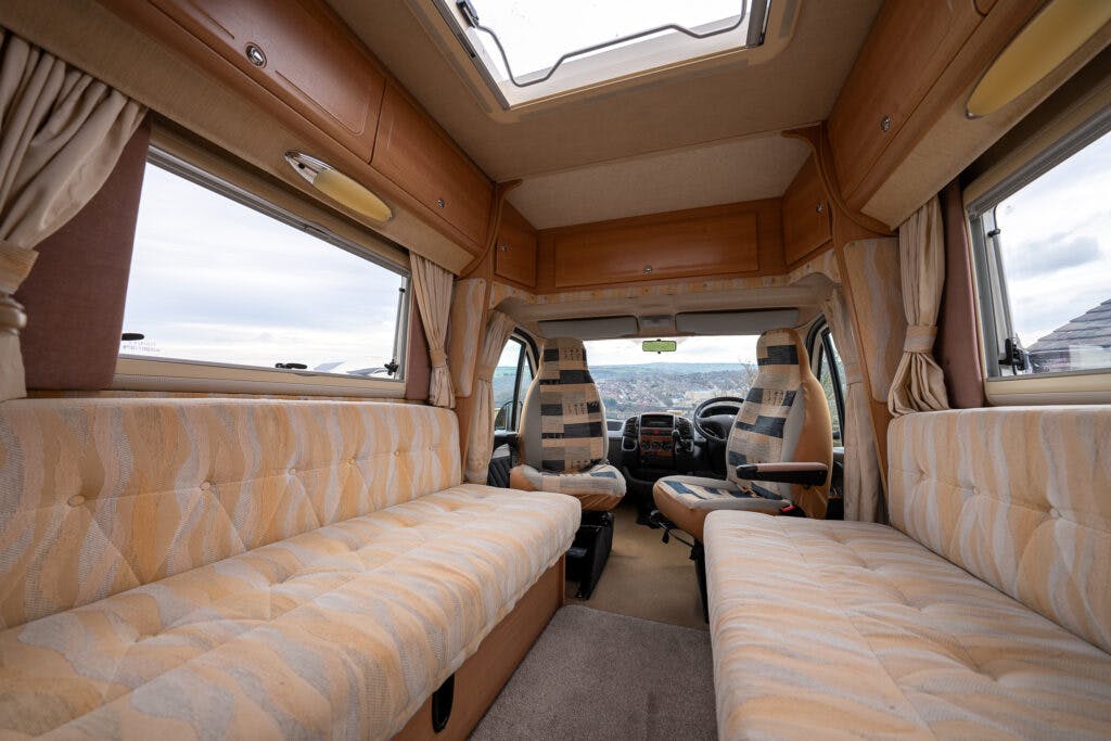 Interior of the 2006 Auto-Sleepers Nuevo EK campervan featuring two cushioned bench seats along the sides, facing each other. The front cab area has two swivel seats. Light wood cabinetry lines the walls and ceiling, and a skylight adds natural light. The windows have beige curtains.