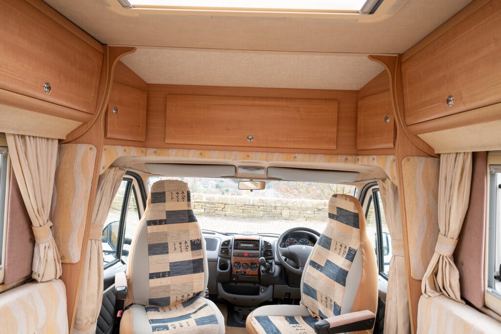 The interior of the 2006 Auto-Sleepers Nuevo EK camper van showcases the driver's seat on the left, a passenger seat on the right, and a control dashboard in the center. Overhead wooden cabinets add to its charm, while striped-pattern upholstery adorns the seats. Curtains hang beside the windows for privacy.