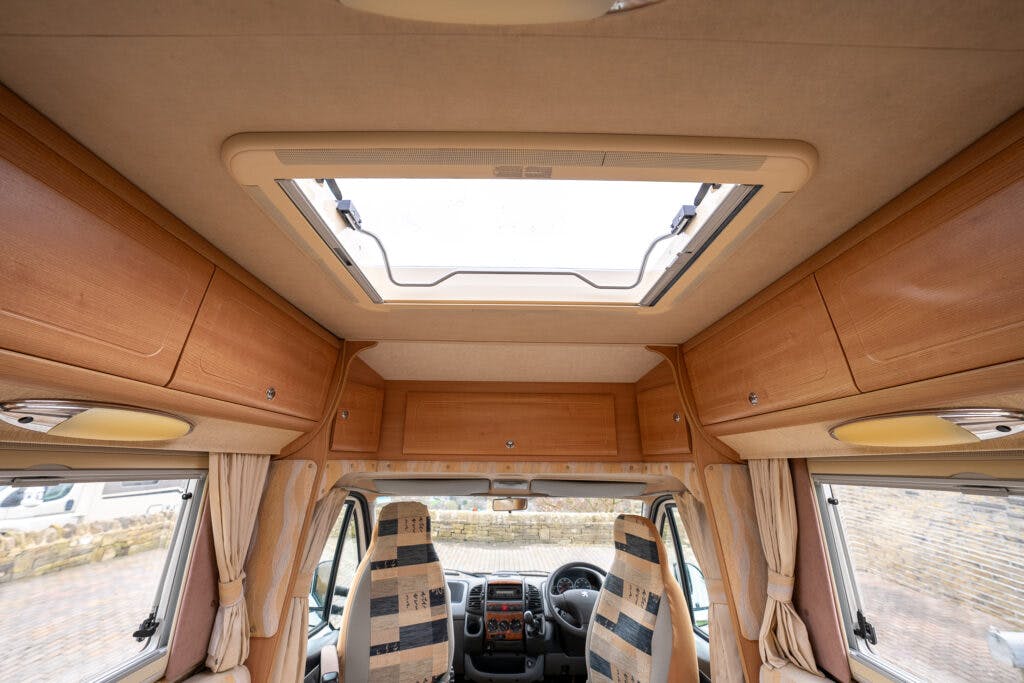 Interior of a 2006 Auto-Sleepers Nuevo EK RV, viewed from the back towards the cockpit. The image shows a ceiling with a skylight and wooden cabinets on both sides. The front windshield and two front seats with patterned upholstery are also visible.