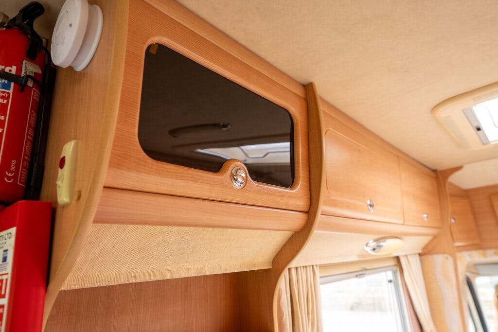 A close-up view of cabinet storage in the 2006 Auto-Sleepers Nuevo EK motorhome. The cabinets are made of light wood with shiny handles. There is a fire extinguisher mounted to the left of the cabinets and a window with curtains below. The beige ceiling adds warmth to the space.