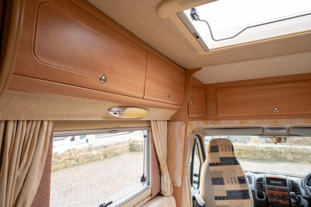 Interior view of a 2006 Auto-Sleepers Nuevo EK camper van showcasing wooden overhead cabinets, a small window with a beige curtain, and a portion of the dashboard. The ceiling features a skylight and the cabinet doors have metallic knobs. Part of a stone wall is visible outside the window.