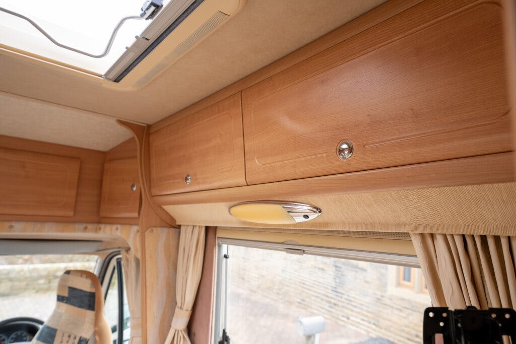 Interior view of a 2006 Auto-Sleepers Nuevo EK motorhome showing wooden overhead cabinets, a window with beige curtains below, and a driver seat partially visible to the left. A ceiling light fixture is also visible near the center of the image.
