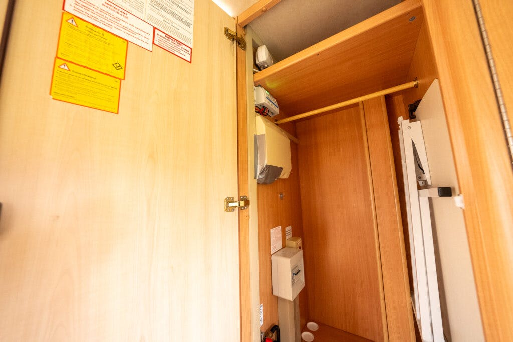 Open wooden closet door revealing inside shelves and compartments, including various electrical components and a small fuse box. Various instruction and warning labels are visible on the closet wall, maintaining the meticulous design reminiscent of the 2006 Auto-Sleepers Nuevo EK.