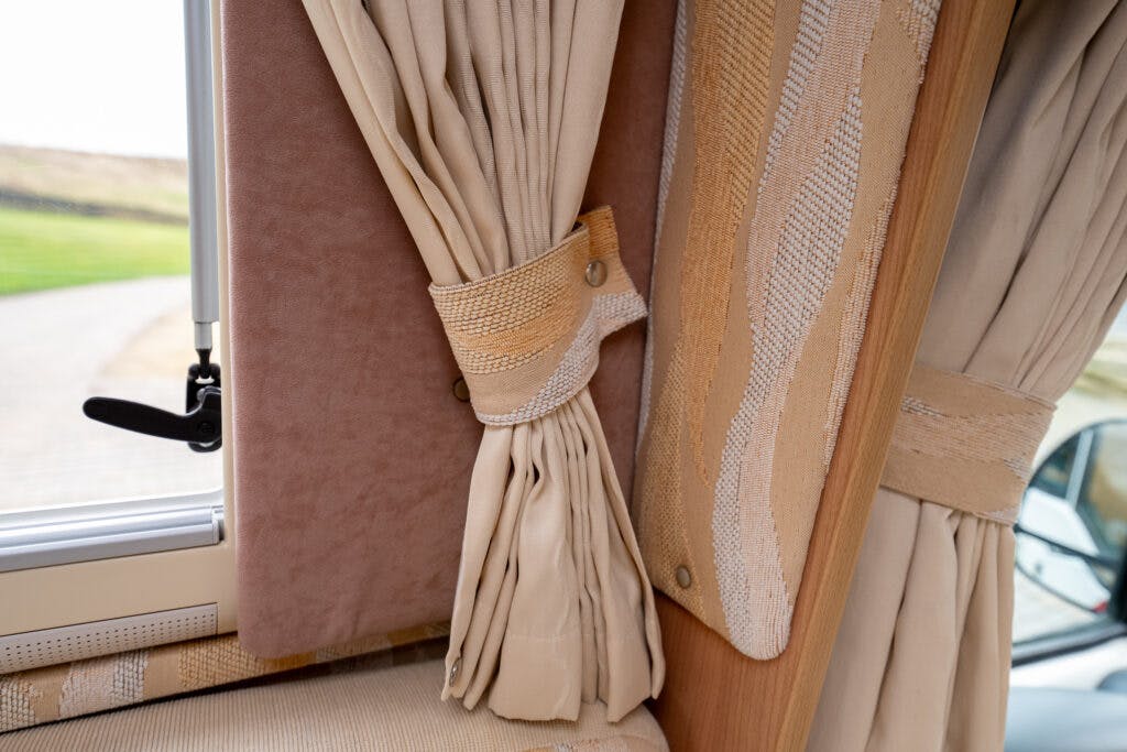 A close-up image of beige curtains in an interior setting, tied back with a matching strap secured by two buttons. The curtain is beside a window with a visible handle, and there is an outside landscape with a path and grassy area, reminiscent of the cozy living space found in a 2006 Auto-Sleepers Nuevo EK.