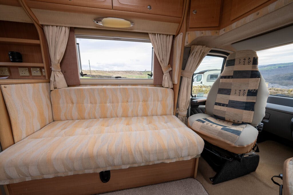 The image shows the interior of a 2006 Auto-Sleepers Nuevo EK camper van with a beige fabric couch and a driver's seat covered in patterned fabric. A large window with curtains is behind the couch, revealing a scenic view of a landscape outside. The space appears compact and cozy.