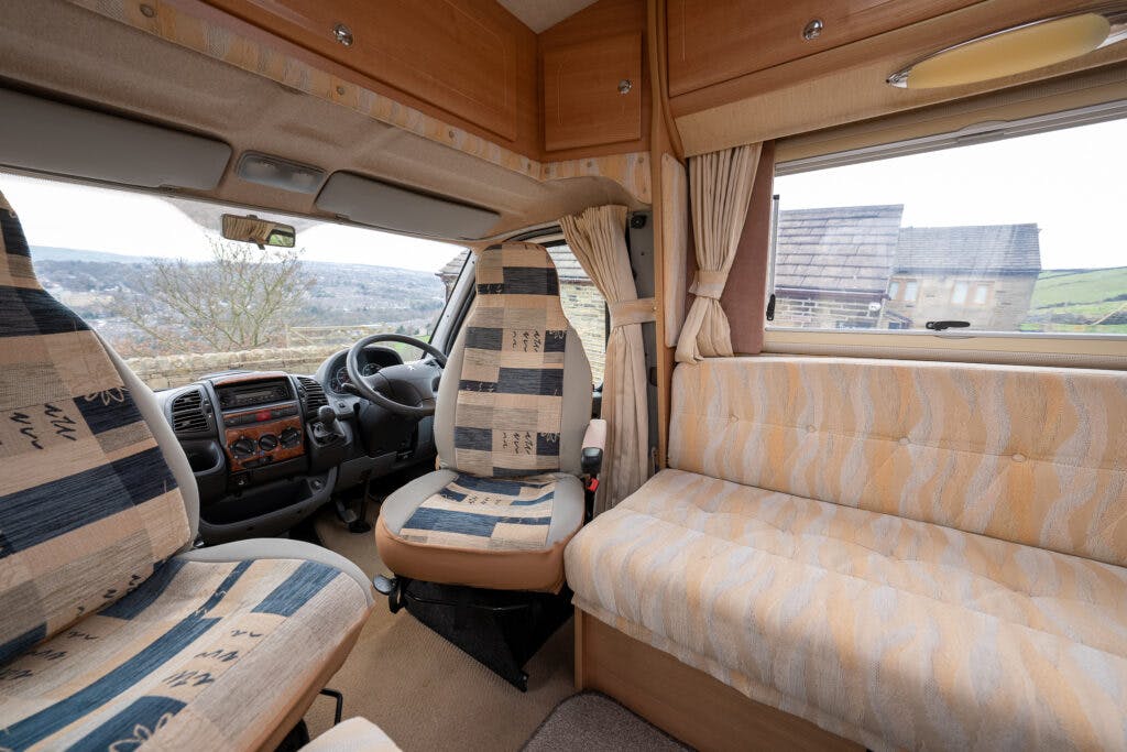 The interior of the 2006 Auto-Sleepers Nuevo EK motorhome shows the front driver's seat and a stationary seat positioned behind it. The seats and couch feature patterned upholstery. A window with curtains reveals a view of a residential area and moorland in the distance.