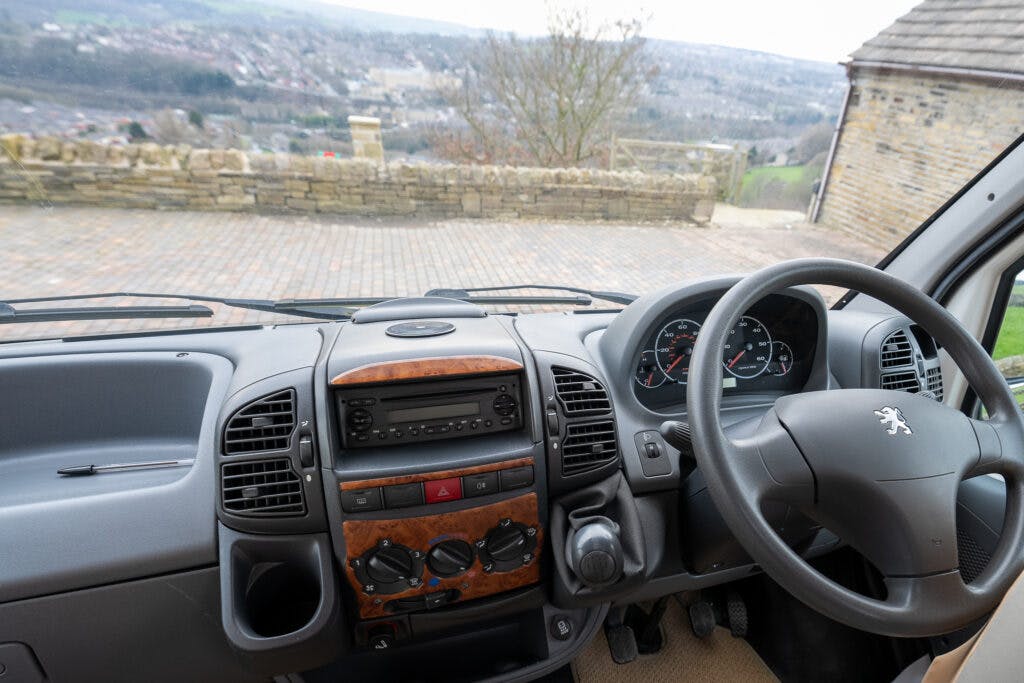 The interior view of the 2006 Auto-Sleepers Nuevo EK's cockpit features a steering wheel, dashboard with a wood finish, various control buttons, air vents, and a radio system. Outside the windshield, a stone fence and a scenic landscape with hills can be seen.