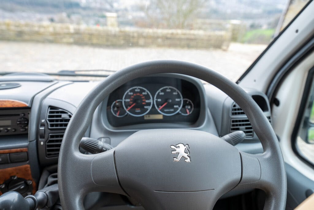 Close-up view from the driver's seat of a Peugeot vehicle, showcasing the steering wheel with the Peugeot lion logo, dashboard gauges, speedometer, and air vents. In the background of this sleek 2006 Auto-Sleepers Nuevo EK, you can see a blurred outdoor scene with stone walls and greenery.