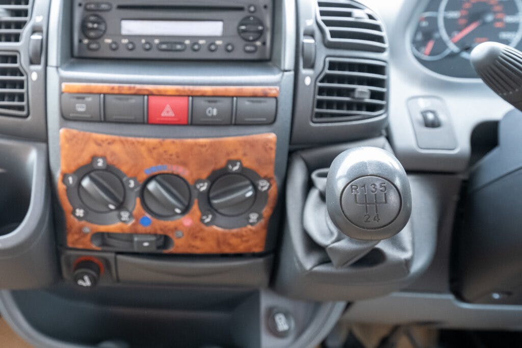 The interior of a 2006 Auto-Sleepers Nuevo EK showcasing the dashboard and center console. The dashboard features a CD player, air conditioning controls, and elegant wooden paneling. A manual gear shift labeled "R13524" is prominently shown to the right, while the speedometer is blurred in the background.