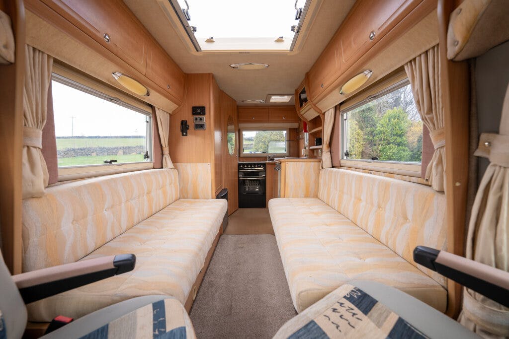 The interior of a beige-colored 2006 Auto-Sleepers Nuevo EK RV features two long, parallel seating areas with patterned cushions facing each other. A compact kitchen area with a stove and small storage compartments is visible at the back. Windows flanking the sides provide natural light.