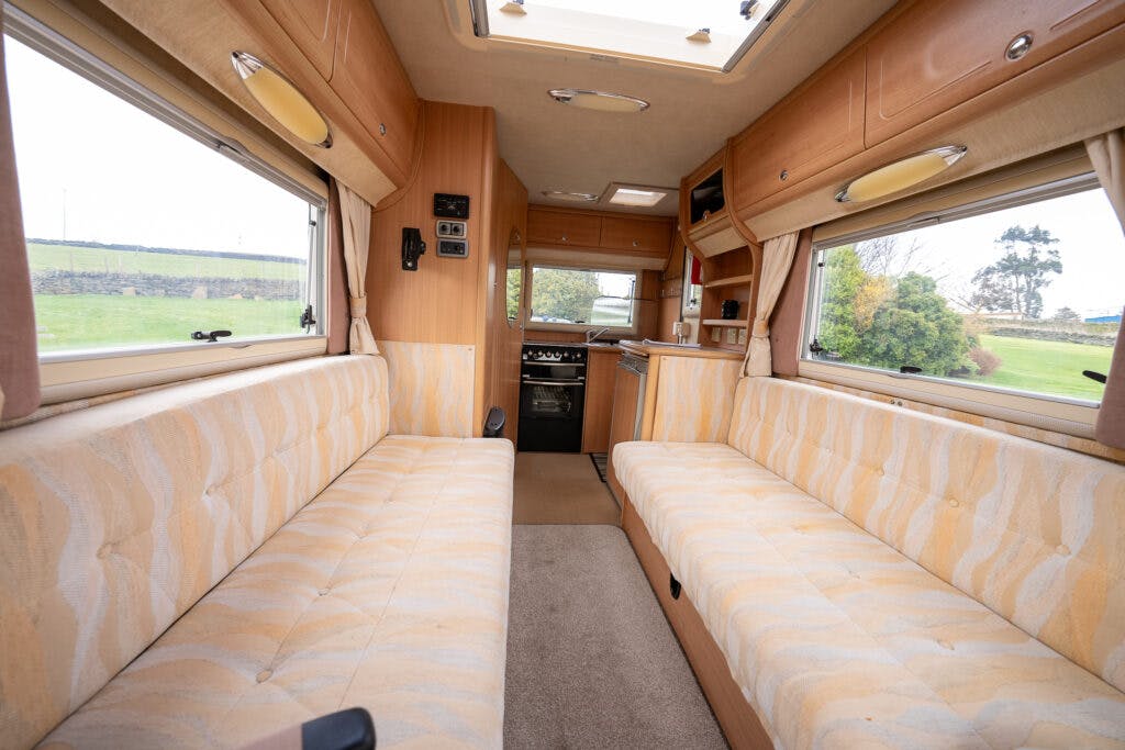 The interior of the 2006 Auto-Sleepers Nuevo EK motorhome features two long, upholstered sofas facing each other, with light-colored patterns. The compact kitchen area includes a stove, sink, and storage cabinets. Windows on both sides provide ample natural light, and a skylight is visible above.