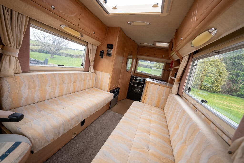 The interior of the 2006 Auto-Sleepers Nuevo EK motorhome features two long, cushioned benches on either side, beige cabinetry, and carpeted flooring. Windows with curtains are on both sides, and a small kitchen area with a stovetop and sink is visible at the far end.