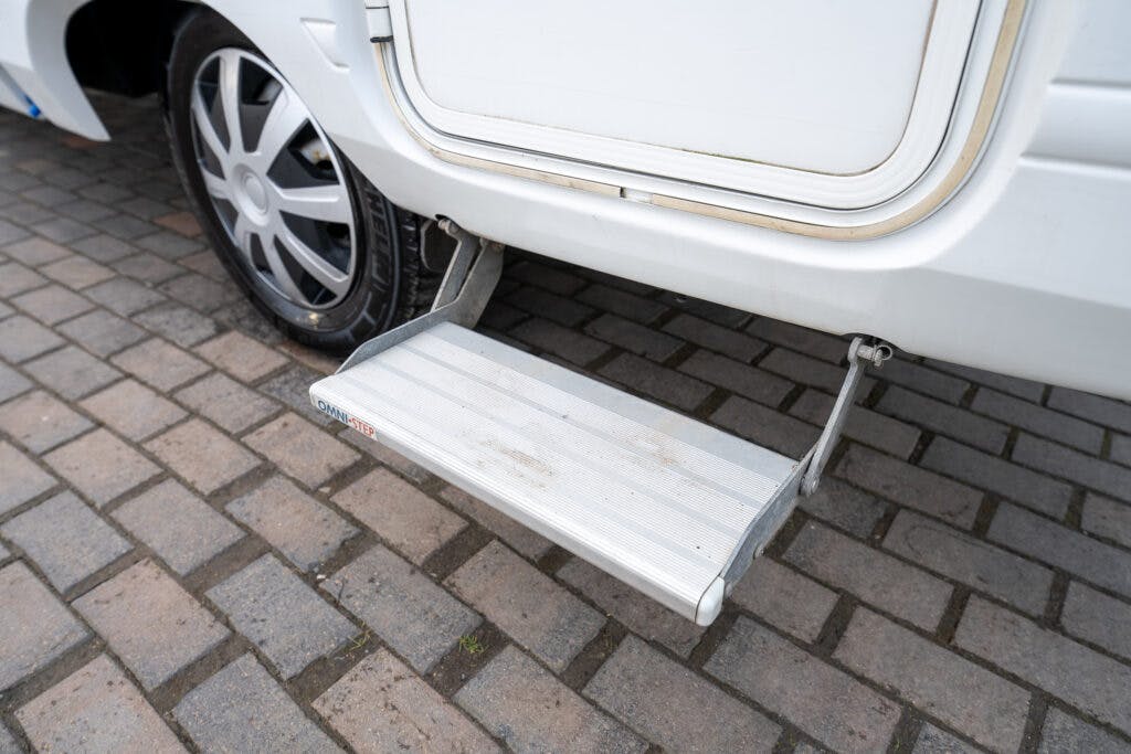 The image shows a metal step attached to the side of a white 2006 Auto-Sleepers Nuevo EK, likely an RV or camper. The step is extended downward to provide easier access to the vehicle's interior. The vehicle is parked on a paved surface made of interlocking bricks.