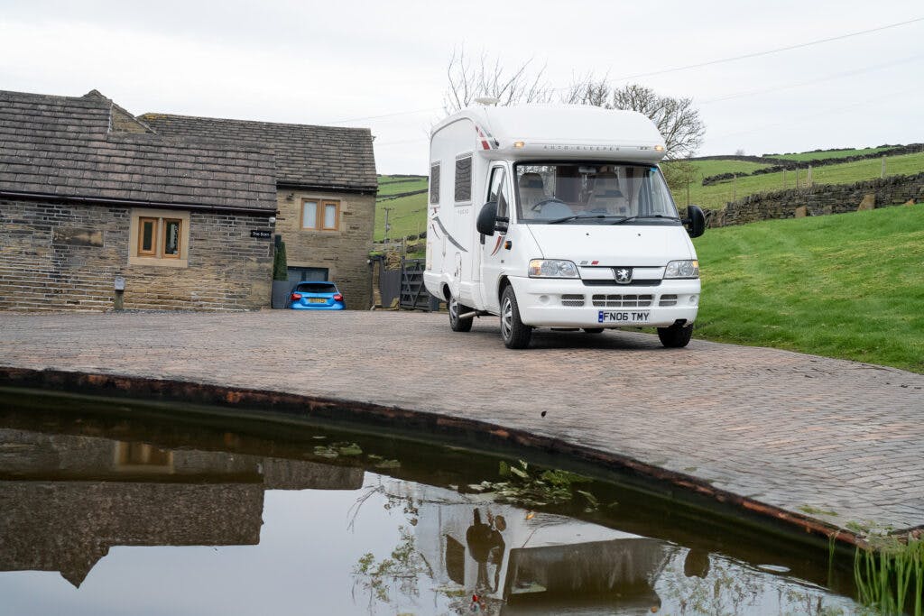 A 2006 Auto-Sleepers Nuevo EK motorhome is parked on a paved driveway near a brick house with a stone roof. A blue car is partially visible behind the house. In the foreground, there is a small pond reflecting the scene, and the background features a grassy hillside.