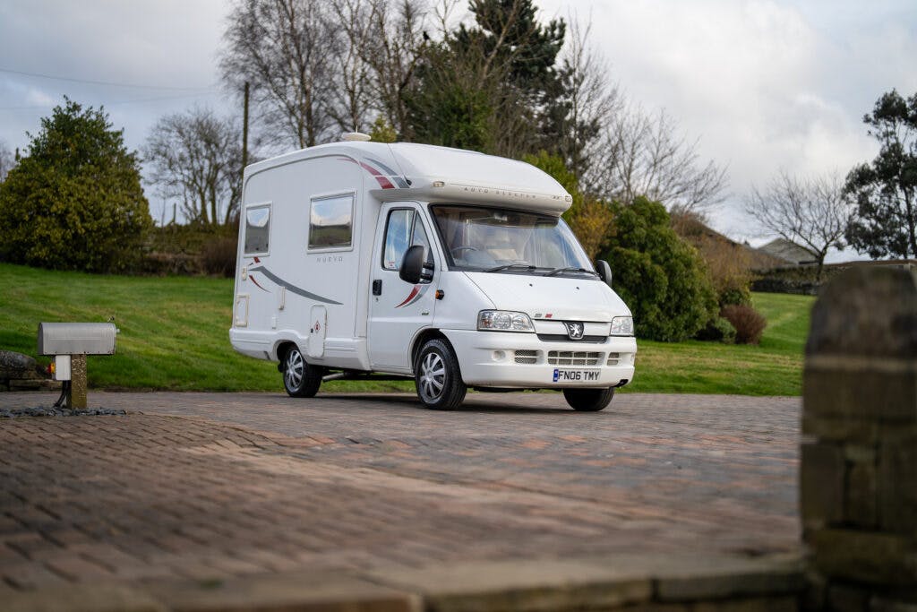 A white 2006 Auto-Sleepers Nuevo EK motorhome is parked on a brick driveway with green grass, trees, and bushes in the background. The motorhome has a Fiat emblem on the front and is positioned at an angle, showcasing its side and front. The sky is overcast with scattered clouds.