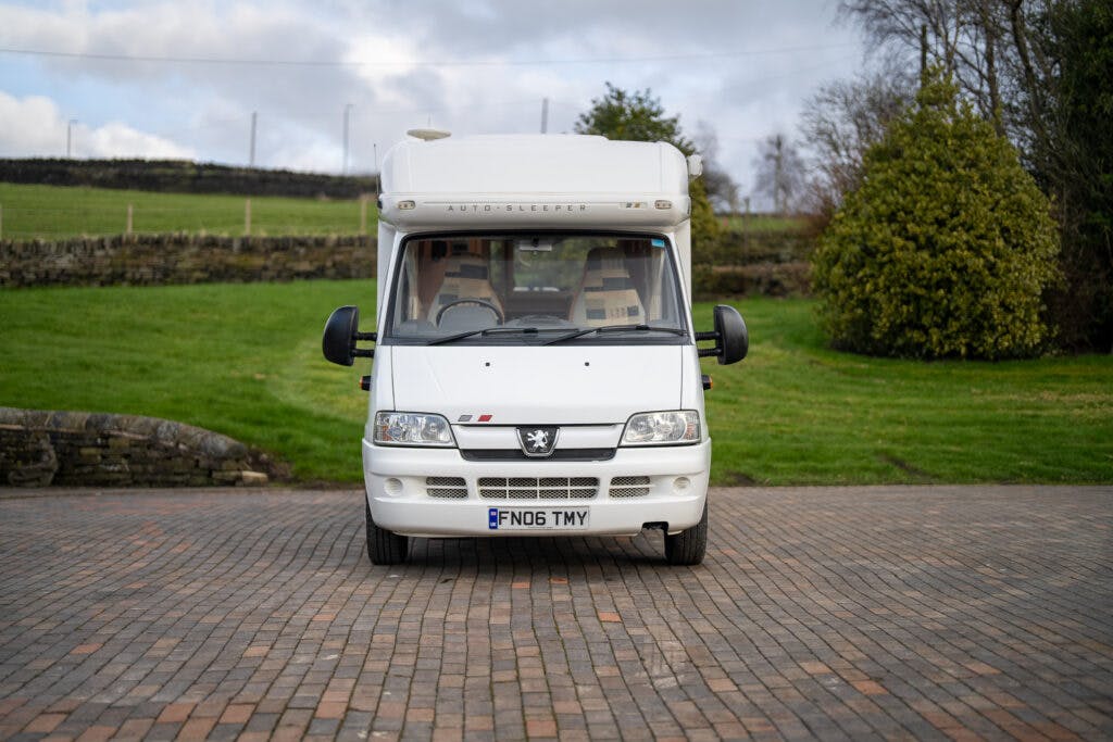A white 2006 Auto-Sleepers Nuevo EK motorhome is parked on a brick driveway with a grass lawn and dry stone wall in the background. The vehicle, bearing the registration "BF06 TMY," is surrounded by a rural environment with trees and a cloudy sky.