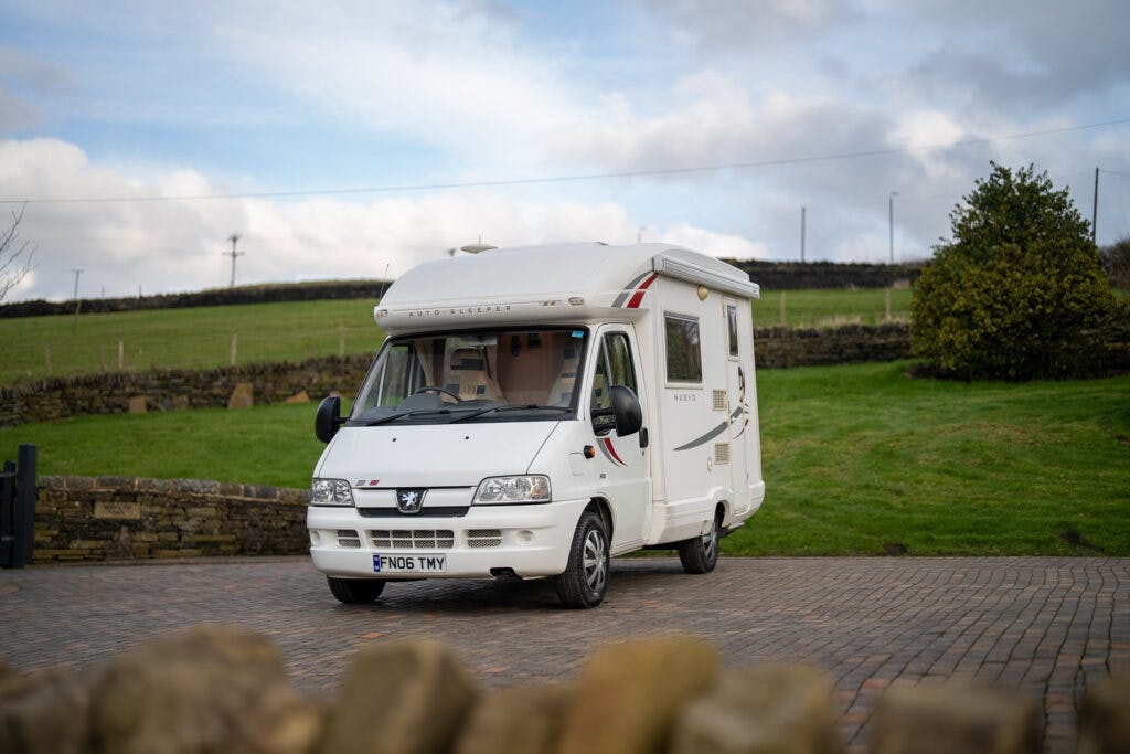 A white 2006 Auto-Sleepers Nuevo EK motorhome is parked on a stone-paved area with green grass and a stone wall in the background. The license plate reads "FN06 TNW." The sky is partly cloudy.