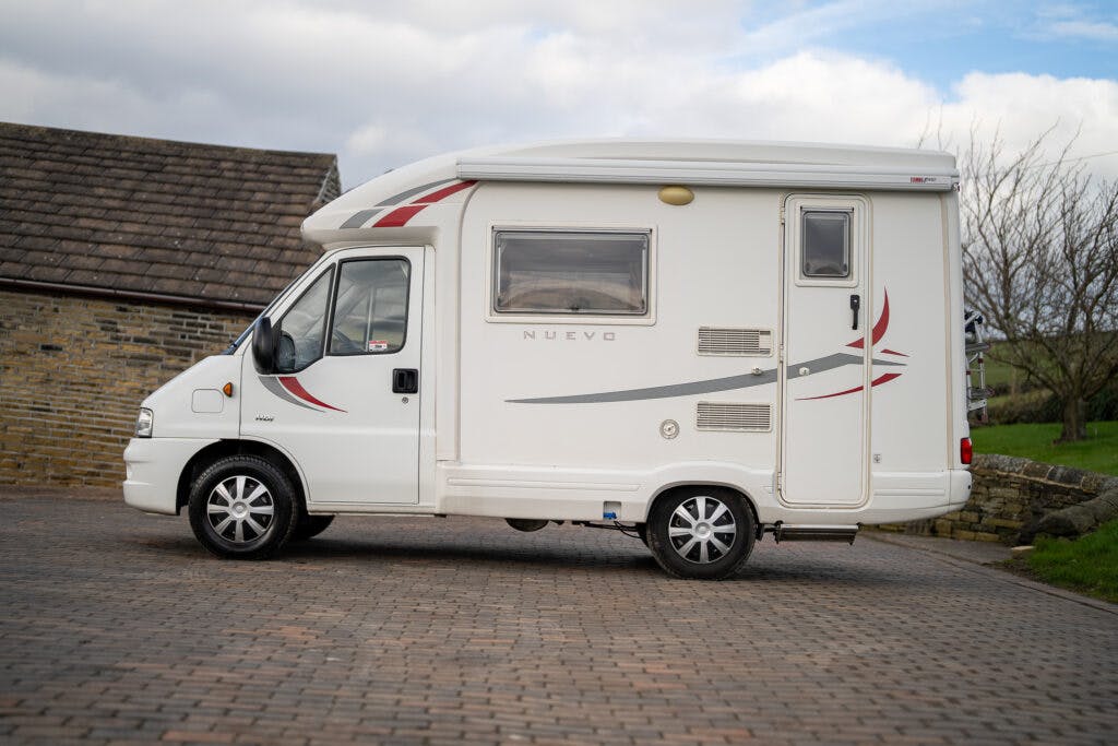 A white 2006 Auto-Sleepers Nuevo EK motorhome is parked on a paved driveway. The vehicle features a single window on the side with red and gray decorative stripes. In the background, there is a portion of a brick building and a grassy area.