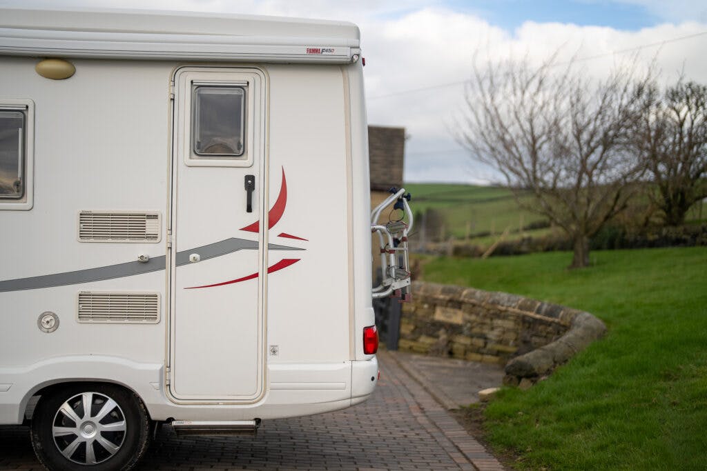 This image shows the rear side of a parked white 2006 Auto-Sleepers Nuevo EK motorhome on a brick driveway. The motorhome has a bike rack attached to its back and is situated in a countryside setting with a stone wall, grassy yard, and trees. The sky is partly cloudy.