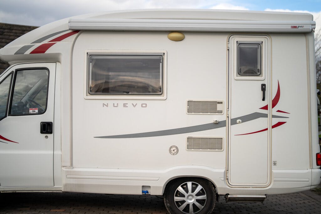 Side view of a white 2006 Auto-Sleepers Nuevo EK motorhome parked on a cobblestone surface. The motorhome features a window, door, and various vents. The word "Nuevo" is visible on the side. Red and grey decals decorate the exterior. The wheel is partially visible at the bottom.
