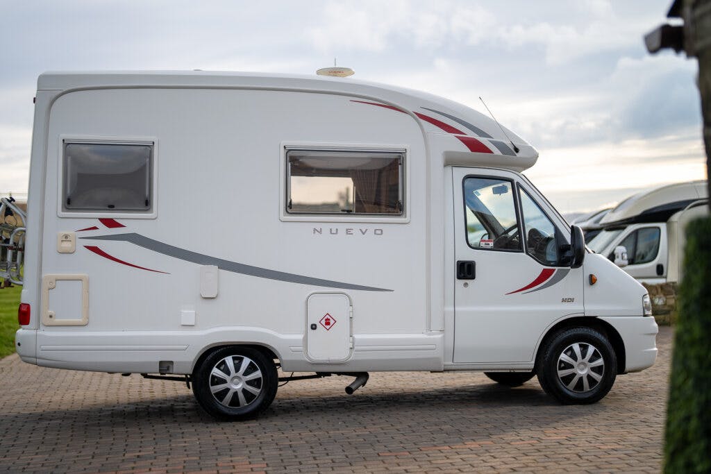 A white, compact motorhome with gray and red accents, labeled "Nuevo" on the side, parked on a cobblestone surface. The 2006 Auto-Sleepers Nuevo EK features two windows on the side and one on the door, with several other motorhomes visible in the background.