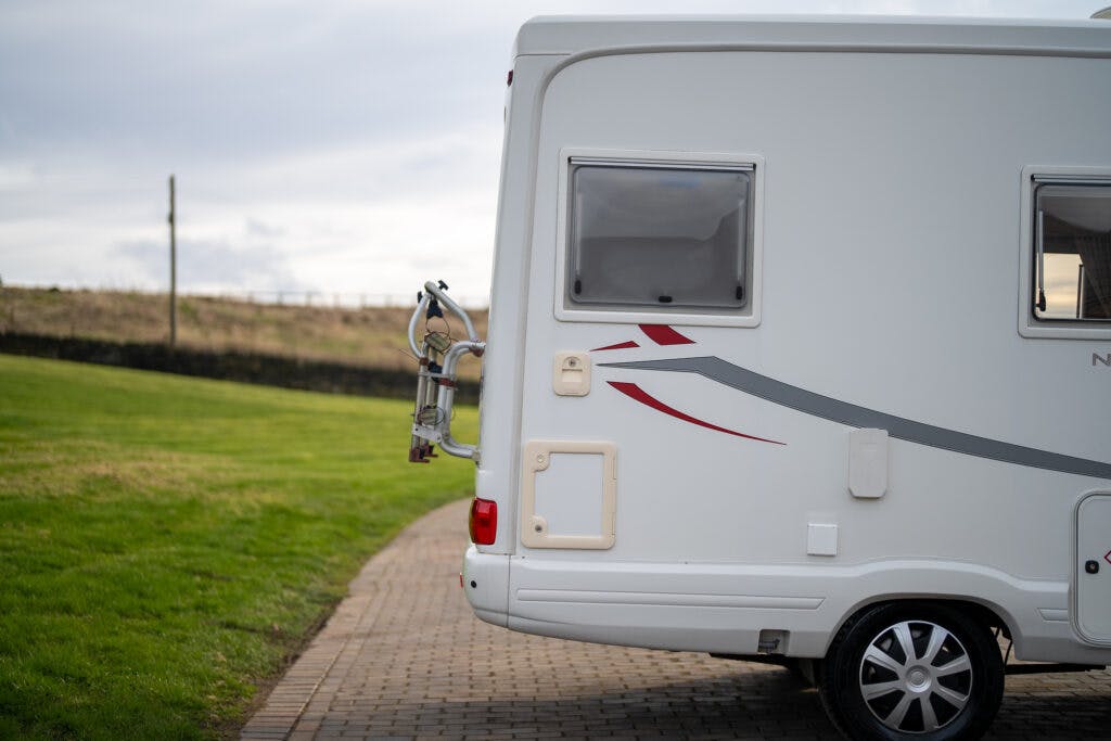 A close-up of the back of a white 2006 Auto-Sleepers Nuevo EK campervan parked on a paved area with a grassy field in the background. The rear bike rack is empty, and two windows are visible on the side of the vehicle. The sky is cloudy.