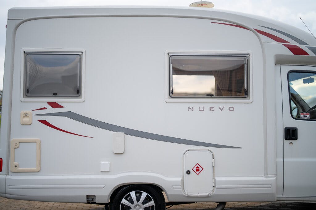Side view of a white 2006 Auto-Sleepers Nuevo EK motorhome parked on a paved surface. The motorhome features two windows, red and grey accent stripes, and storage compartments. The wheel and part of the driver's side door are visible.