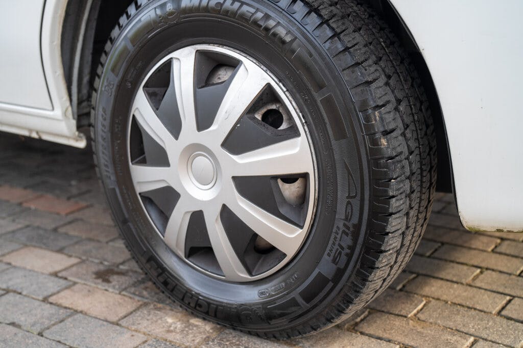 Close-up of a 2006 Auto-Sleepers Nuevo EK's front right wheel on a paved driveway. The wheel features a black Michelin tire with light tread wear and a silver hubcap with a multi-spoke design. The white car body is partially visible.