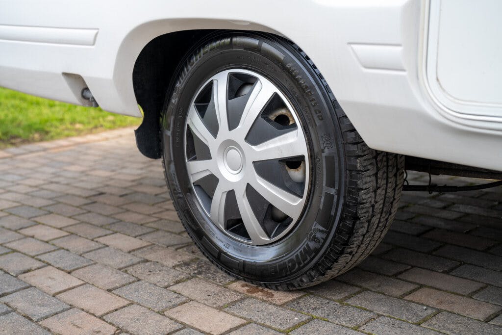 Close-up of the rear wheel of a white 2006 Auto-Sleepers Nuevo EK, parked on a brick driveway. The tire appears to be in good condition with a visible tread pattern. The alloy wheel has a multi-spoke design, and grass can be seen in the background.
