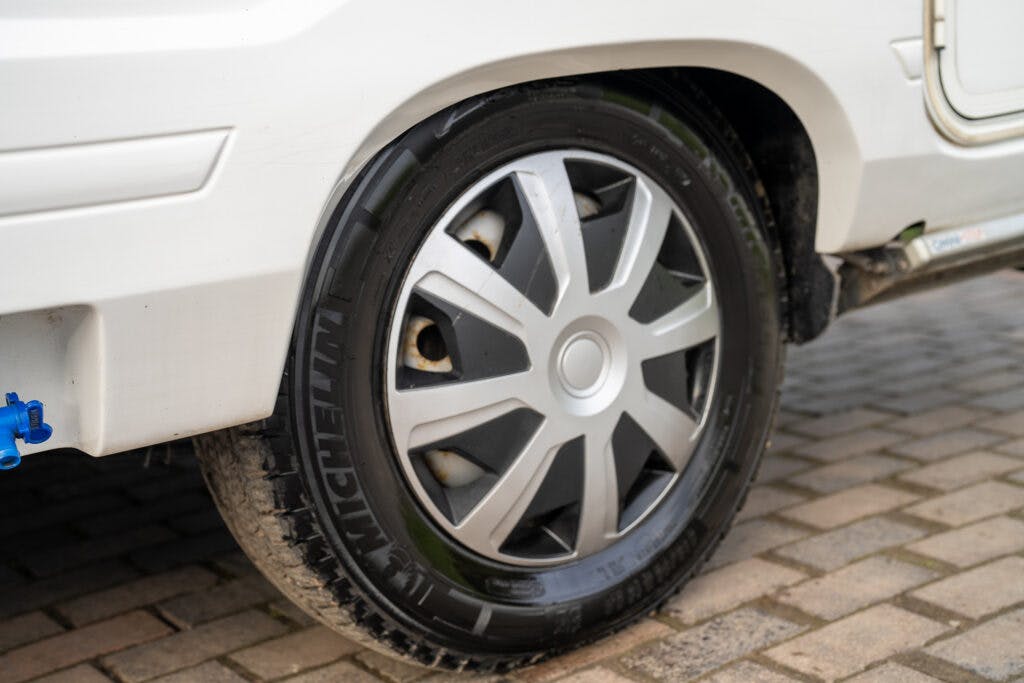 A close-up view of the rear wheel of a white 2006 Auto-Sleepers Nuevo EK, featuring a Michelin tire and a silver alloy wheel. The vehicle is parked on a paved surface with interlocking bricks. There is a small blue object attached to the wheel arch.