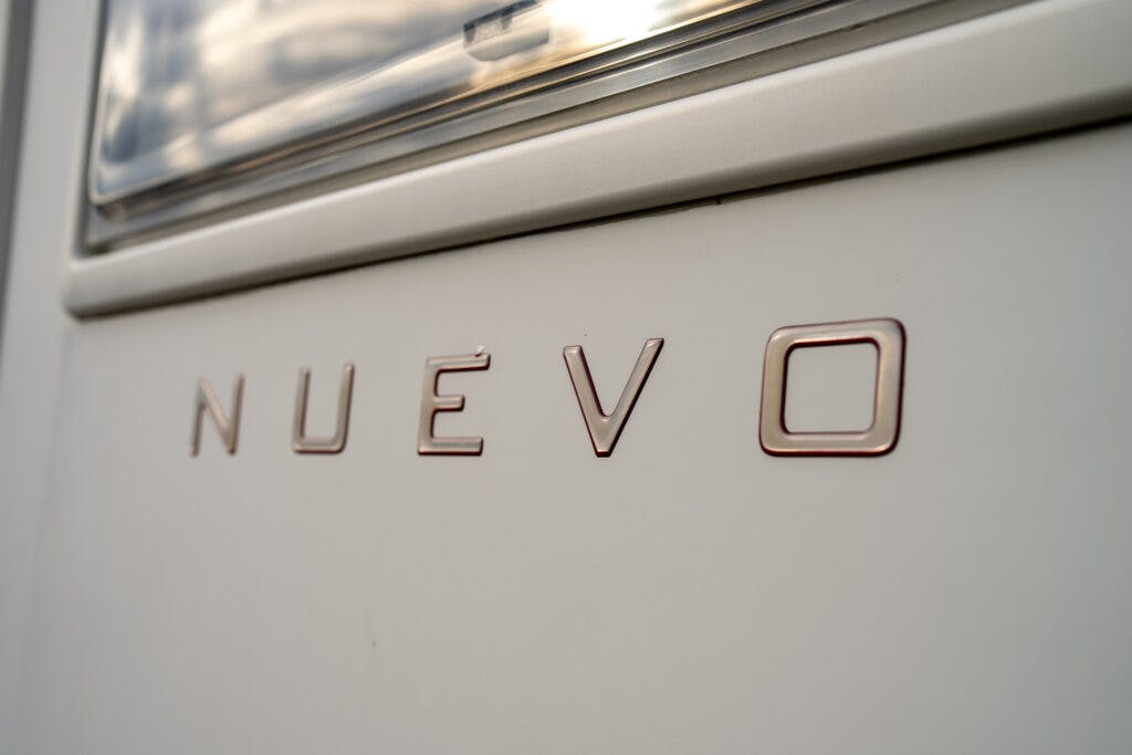 Close-up shot of a metallic surface displaying the word "NUEVO" in raised letters. The background is part of a 2006 Auto-Sleepers Nuevo EK, with a reflective surface above the text. The letters have a slight shine and a modern design.